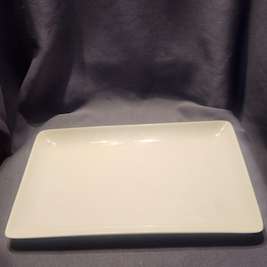 以下が含まれることがあります： 長方形の白いセラミック製サービングプラッター。プラッターはわずかに盛り上がった縁と滑らかで光沢のある表面を持っています。その形状はモダンでミニマリストで、前菜やデザートの提供に適しています。