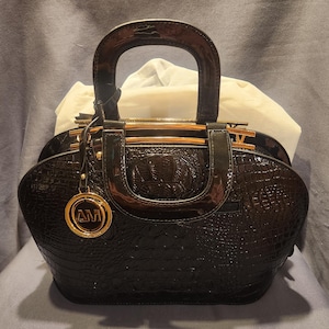 Puede incluir: Un bolso negro brillante con textura de piel de cocodrilo. Cuenta con un asa curva, detalles dorados y un charm circular con las letras "AM". El interior está relleno de tela blanca.
