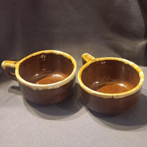 Può includere: Due ciotole di zuppa in ceramica marrone con manici. Le ciotole hanno una finitura lucida e un bordo color crema. L'interno delle ciotole è marrone più scuro. Le ciotole sono vuote e poggiano su una superficie grigia.
