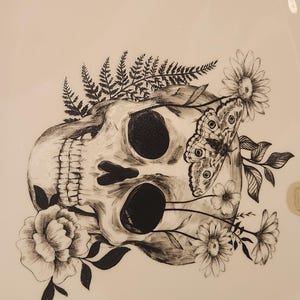 Puede incluir: Ilustración en blanco y negro de una calavera adornada con flores, helechos y una mariposa. Las cuencas de los ojos y la cavidad nasal de la calavera son negras. Las flores incluyen margaritas y una peonía. La mariposa tiene un patrón detallado en sus alas.