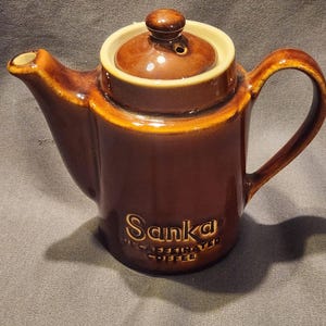 May include: A brown ceramic teapot with a matching lid. The teapot has a curved handle and spout, and the words "Sanka Decaffeinated Coffee" are printed on the front. The rim of the teapot and lid are a lighter tan color.