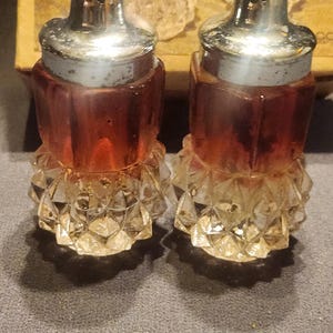 Puede incluir: Un par de saleros y pimenteros antiguos. Los saleros y pimenteros tienen una base transparente facetada y un cuerpo facetado de color marrón rojizo. Están rematados con una tapa de metal plateado. Los saleros y pimenteros son probablemente de vidrio.