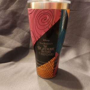 May include: A tapered tumbler featuring a graphic design inspired by The Nightmare Before Christmas, with sections of pink, black, and blue patterns. The text Disney Tim Burton's The Nightmare Before Christmas is printed in the center.