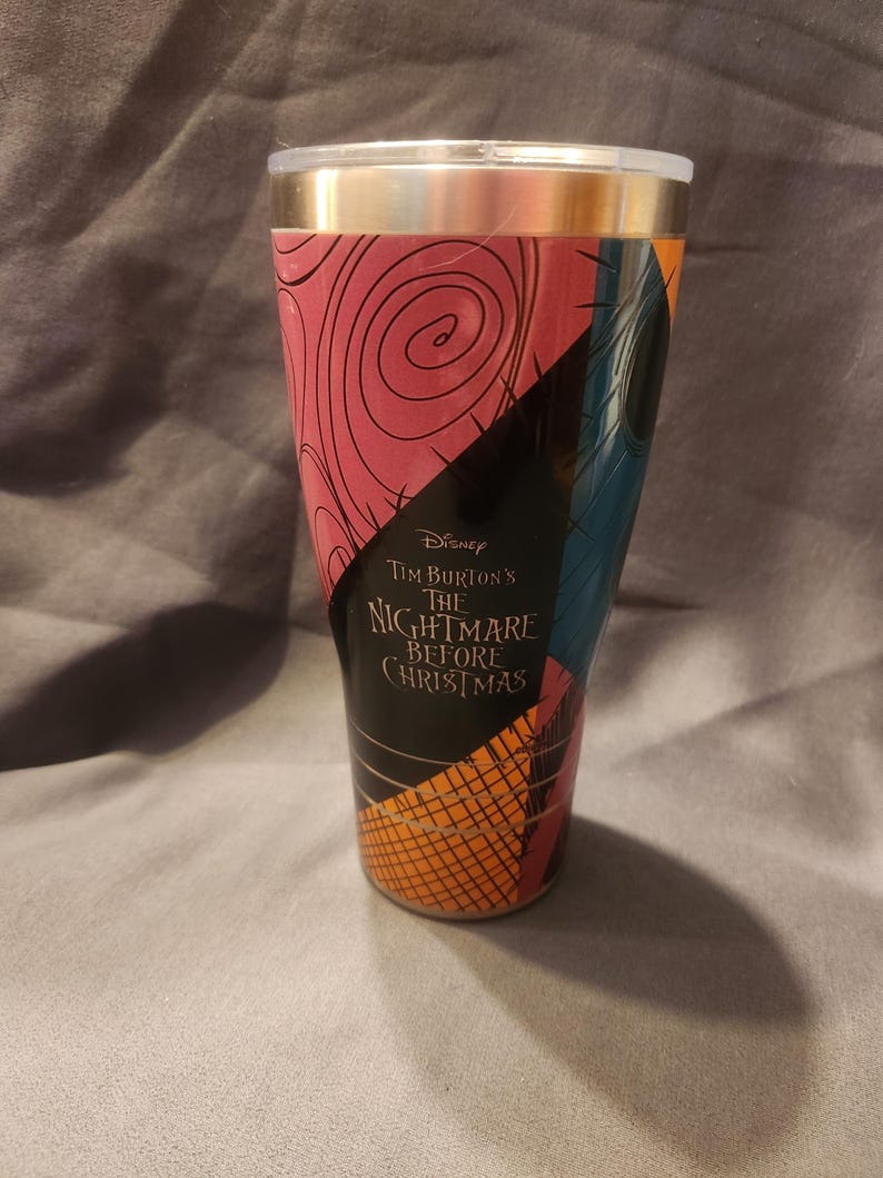 May include: A tapered tumbler featuring a graphic design inspired by The Nightmare Before Christmas, with sections of pink, black, and blue patterns. The text Disney Tim Burton's The Nightmare Before Christmas is printed in the center.