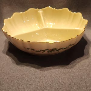 Può includere: Un piatto da portata color crema a tre scomparti con bordo smerlato e finiture dorate. Il piatto presenta un motivo di agrifoglio e bacche attorno alla base. Il piatto è probabilmente in porcellana o ceramica, adatto per snack.