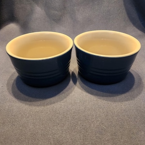 Può includere: Due pirofile in ceramica blu navy con interno color crema. Le pirofile hanno un esterno a coste e sono riempite con un liquido trasparente. Le pirofile sono su una superficie blu.