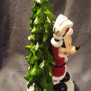 May include: A ceramic Christmas figurine featuring Mickey Mouse dressed as Santa Claus, standing next to a green Christmas tree with white accents. Mickey is wearing a red suit with white trim and a Santa hat. The figurine is on a white base.