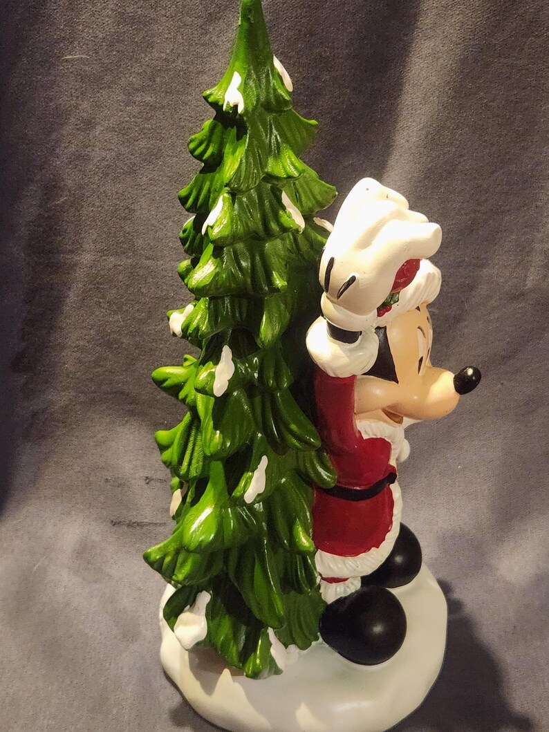 May include: A ceramic Christmas figurine featuring Mickey Mouse dressed as Santa Claus, standing next to a green Christmas tree with white accents. Mickey is wearing a red suit with white trim and a Santa hat. The figurine is on a white base.