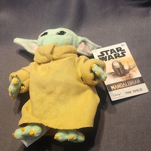 May include: A plush toy of The Child from Star Wars: The Mandalorian. The figure has a light green face and hands, large ears, and is wearing a yellow coat. A Star Wars tag is attached.