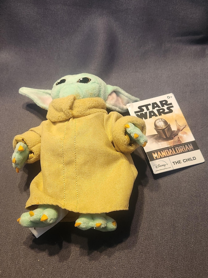 May include: A plush toy of The Child from Star Wars: The Mandalorian. The figure has a light green face and hands, large ears, and is wearing a yellow coat. A Star Wars tag is attached.