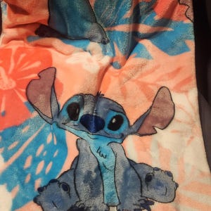 Puede incluir: Una manta de felpa con el personaje Stitch de Lilo & Stitch. La manta tiene un estampado floral rosa, naranja y azul. Stitch est&aacute; representado en azul con orejas grandes y una expresi&oacute;n amigable. La manta es suave y acogedora.