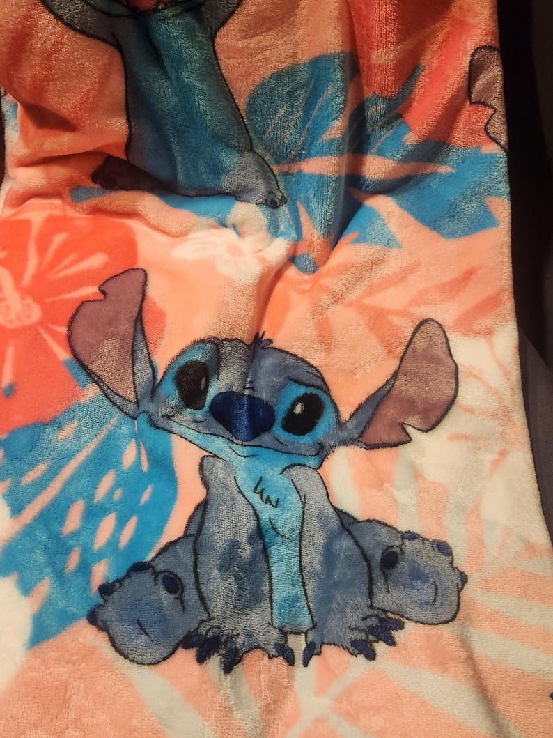 Puede incluir: Una manta de felpa con el personaje Stitch de Lilo & Stitch. La manta tiene un estampado floral rosa, naranja y azul. Stitch est&aacute; representado en azul con orejas grandes y una expresi&oacute;n amigable. La manta es suave y acogedora.