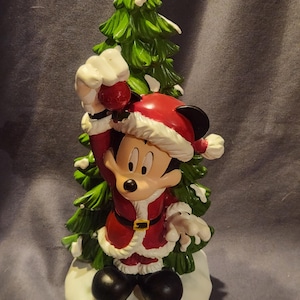 May include: A festive figurine of Mickey Mouse dressed in a Santa Claus outfit, holding a red ornament. The figure stands in front of a green Christmas tree with white accents, on a snowy base. The figurine is a holiday decoration.