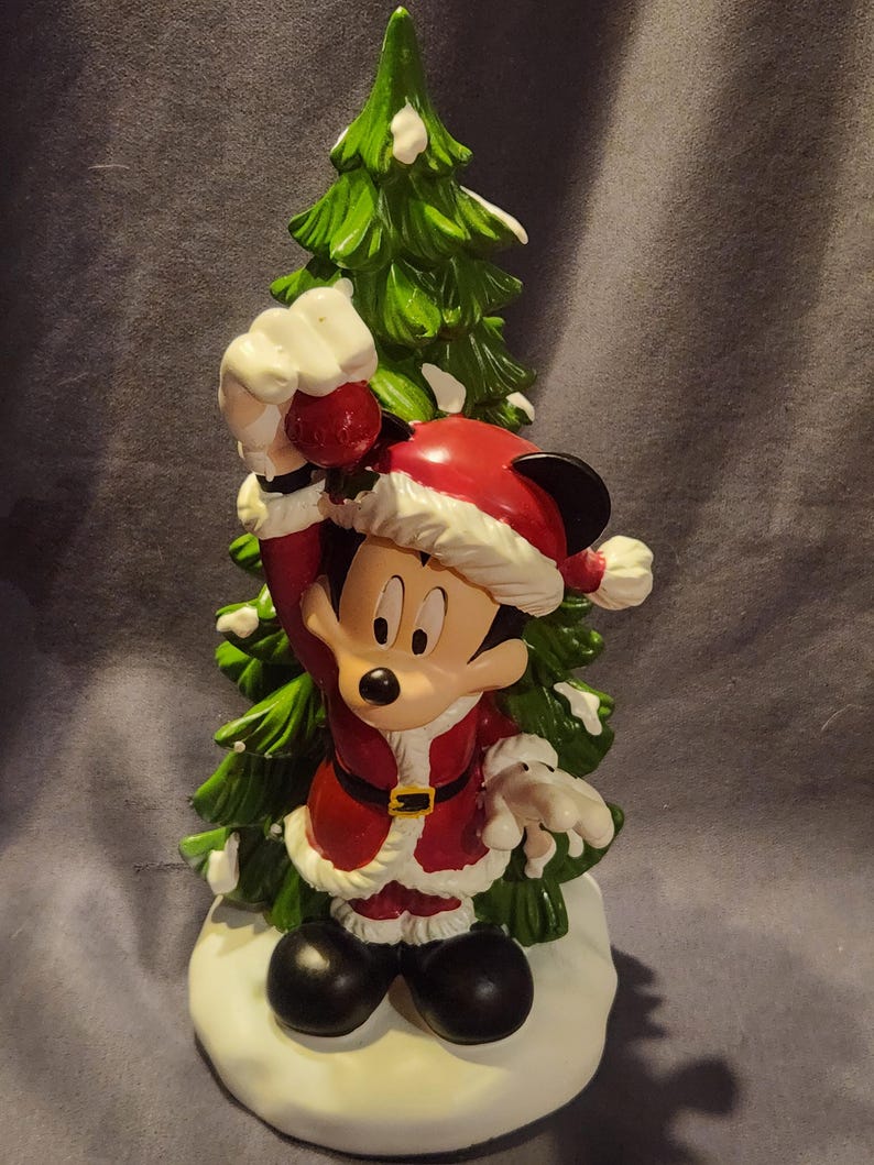 May include: A festive figurine of Mickey Mouse dressed in a Santa Claus outfit, holding a red ornament. The figure stands in front of a green Christmas tree with white accents, on a snowy base. The figurine is a holiday decoration.