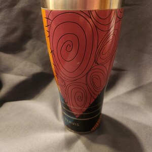 May include: A Tervis stainless steel tumbler featuring a tapered design with a maroon and orange graphic pattern of swirling lines. The cup has a metallic silver rim at the top and a clear plastic lid. The brand name Tervis is printed in white.