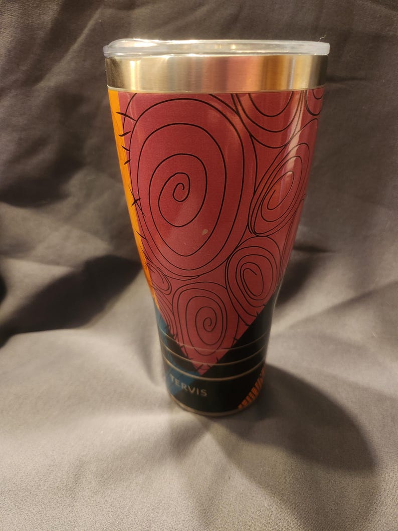 May include: A Tervis stainless steel tumbler featuring a tapered design with a maroon and orange graphic pattern of swirling lines. The cup has a metallic silver rim at the top and a clear plastic lid. The brand name Tervis is printed in white.