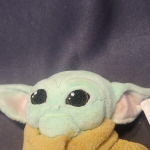 May include: A plush toy of a character with large black eyes, large ears, and a light green complexion. The character is wearing a tan-colored outfit. The toy is a popular character from a science fiction franchise.