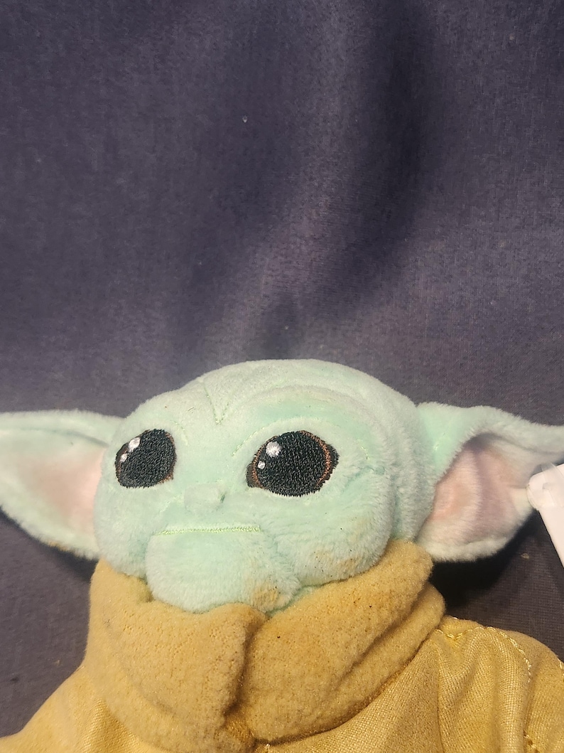 May include: A plush toy of a character with large black eyes, large ears, and a light green complexion. The character is wearing a tan-colored outfit. The toy is a popular character from a science fiction franchise.