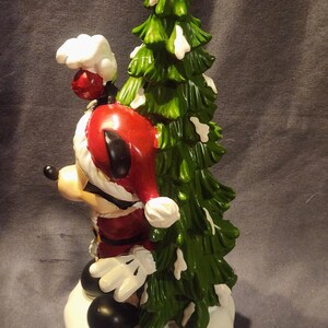 May include: A ceramic figurine of Mickey Mouse dressed as Santa Claus, holding a green Christmas tree with white accents. Mickey is wearing a red Santa hat and suit, white gloves, and black shoes. The figurine is on a white base.