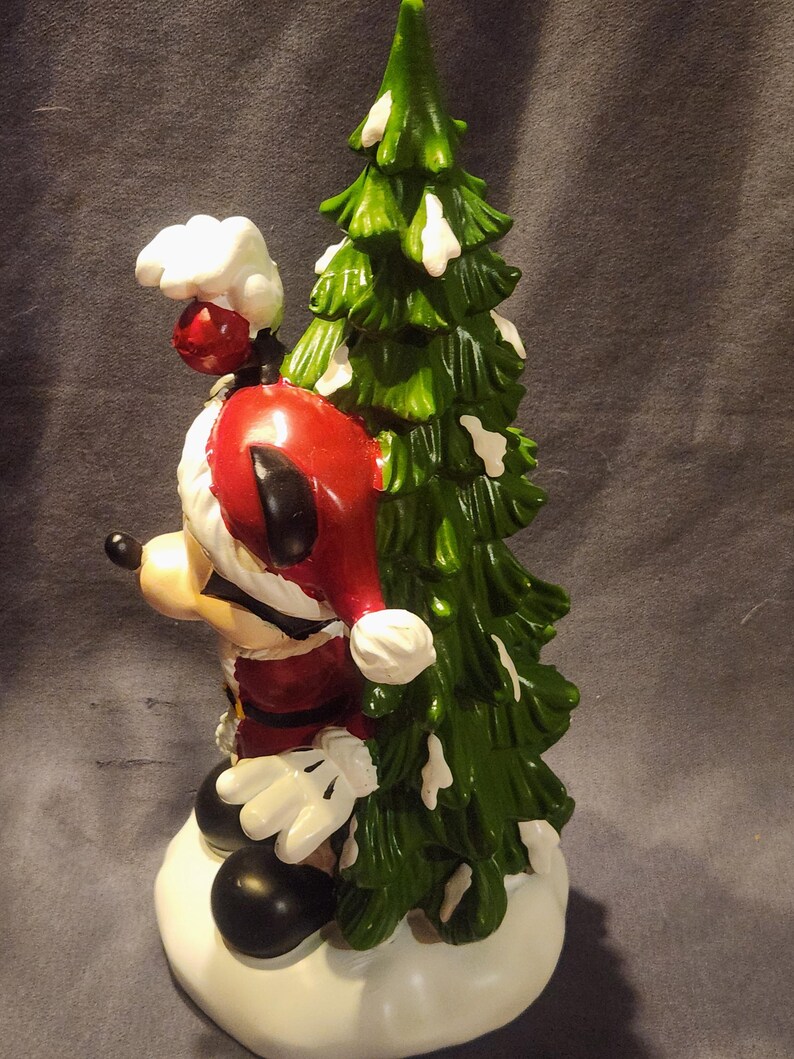 May include: A ceramic figurine of Mickey Mouse dressed as Santa Claus, holding a green Christmas tree with white accents. Mickey is wearing a red Santa hat and suit, white gloves, and black shoes. The figurine is on a white base.