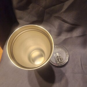 May include: A cylindrical stainless steel tumbler with a metallic interior finish and a circular rim. Beside the open tumbler lies a clear, circular plastic lid featuring a sliding closure mechanism and a small, visible brand logo.