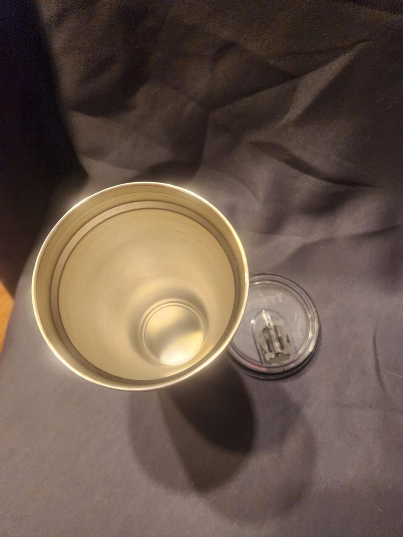 May include: A cylindrical stainless steel tumbler with a metallic interior finish and a circular rim. Beside the open tumbler lies a clear, circular plastic lid featuring a sliding closure mechanism and a small, visible brand logo.