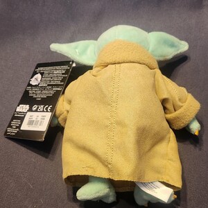 May include: A plush toy of Grogu, also known as Baby Yoda, from the Star Wars series. The toy has large ears, green skin, and is wearing a tan robe. A black tag is attached to the toy.