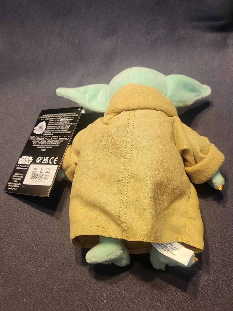 May include: A plush toy of Grogu, also known as Baby Yoda, from the Star Wars series. The toy has large ears, green skin, and is wearing a tan robe. A black tag is attached to the toy.