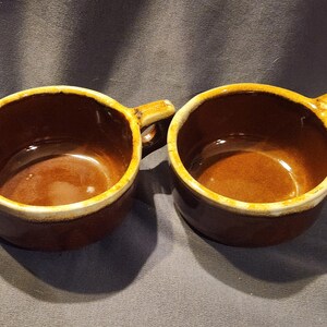 Può includere: Due ciotole da zuppa in ceramica marrone con manici. Le ciotole hanno una finitura lucida e un bordo color crema. L'interno delle ciotole è di una tonalità di marrone più chiara. Le ciotole sono probabilmente vintage o antiche.