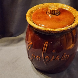 May include: A brown ceramic cookie jar with a lid. The jar has a rounded shape and a glossy finish. The lid is a lighter shade of brown and has a small handle. The word "cookies" is written on the side.