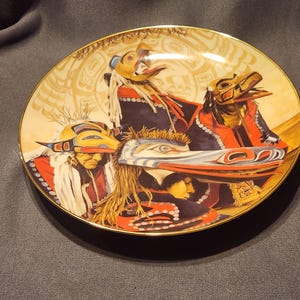 May include: Decorative plate with a Native American design. The plate has a gold rim and depicts figures in ceremonial attire with masks. The artwork includes shades of red, black, and gold, with intricate details.