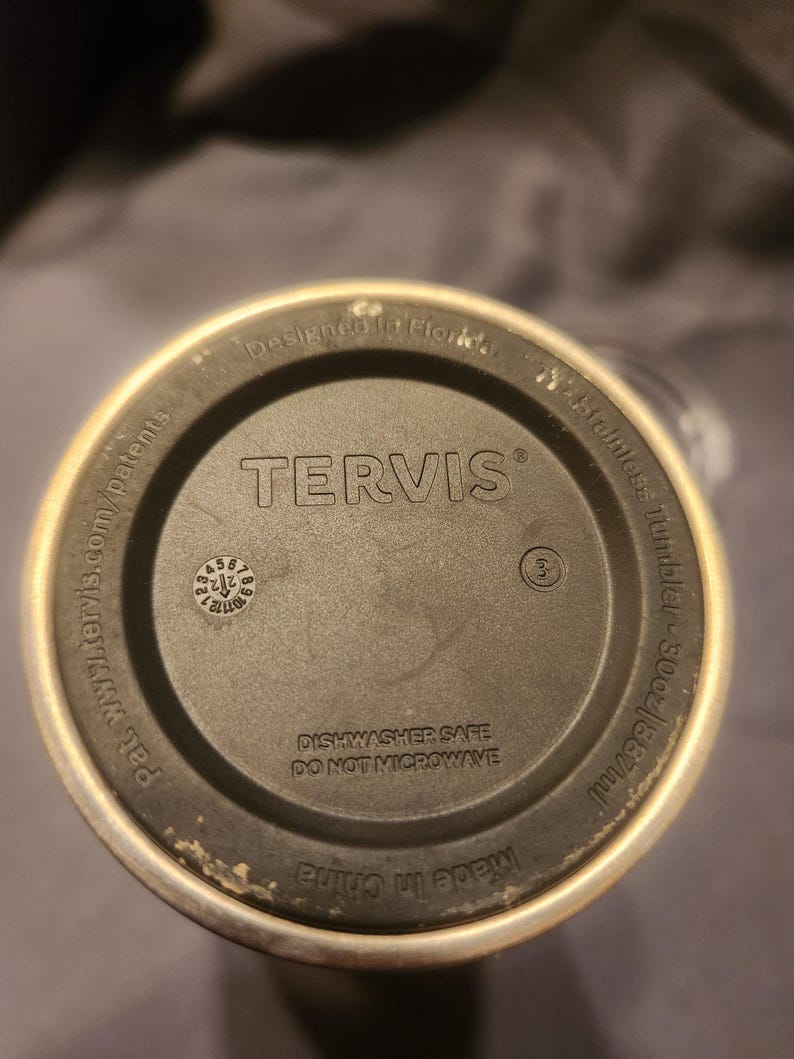 May include: The circular base of a Tervis stainless steel tumbler with a 30oz capacity. The black bottom features the text 'TERVIS', 'Designed in Florida', 'DISHWASHER SAFE', 'DO NOT MICROWAVE', 'Made in China', and a small circular date dial.