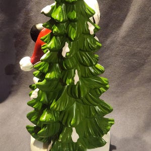 May include: A ceramic Christmas tree figurine with a green, snow-dusted design. The figurine features a red and white Santa hat and a black head. The base is white. The figurine is a Disney product.
