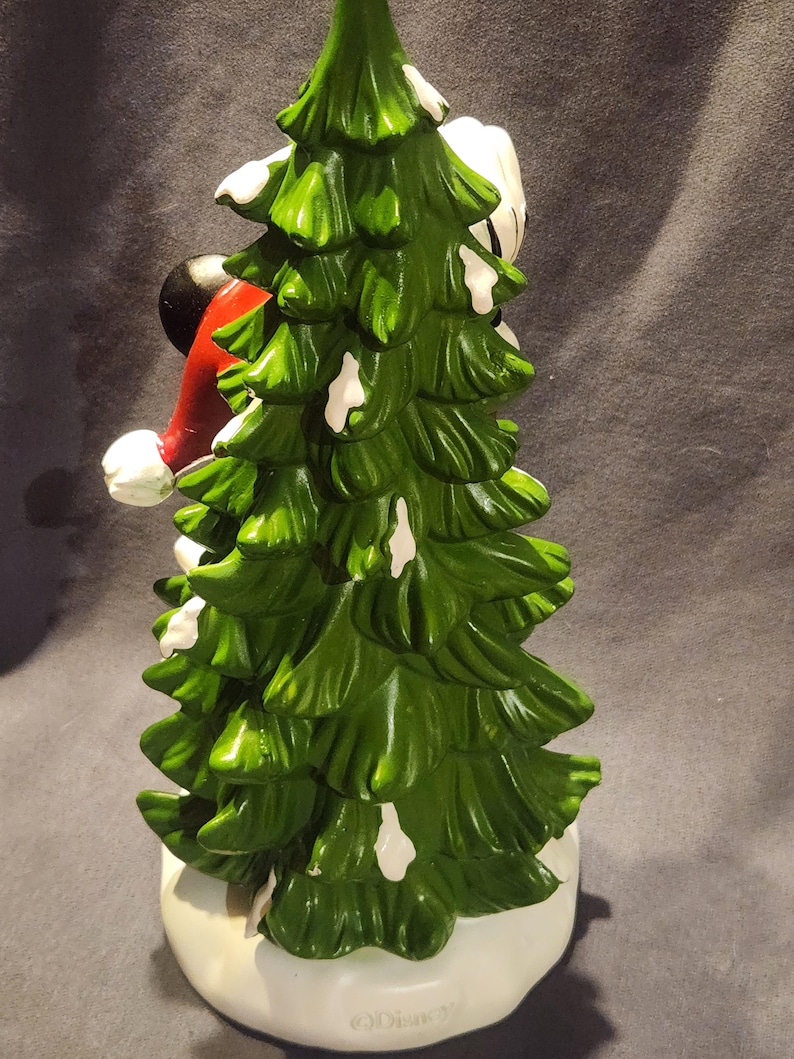 May include: A ceramic Christmas tree figurine with a green, snow-dusted design. The figurine features a red and white Santa hat and a black head. The base is white. The figurine is a Disney product.