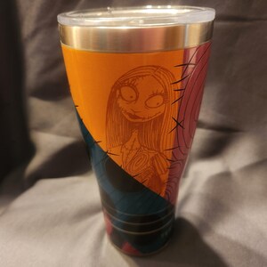 May include: A stainless steel tumbler featuring a graphic print of the character Sally from The Nightmare Before Christmas. The design includes orange, blue, and pink geometric sections with black stitched detailing and a clear plastic lid.
