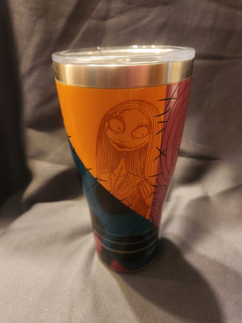 May include: A stainless steel tumbler featuring a graphic print of the character Sally from The Nightmare Before Christmas. The design includes orange, blue, and pink geometric sections with black stitched detailing and a clear plastic lid.