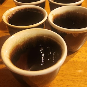 Może przedstawiać: Pięć brązowych ceramicznych kubków z białym rantem i brązową glazurą wewnątrz. Kubki są wypełnione ciemnym płynem, prawdopodobnie kawą lub herbatą.