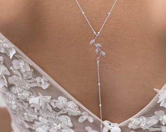 May include: A delicate silver necklace with a leaf design and a dangling pearl pendant. The necklace features small, round beads along the chain and is worn on a white, floral-embroidered dress. The necklace is a bridal or special occasion accessory.
