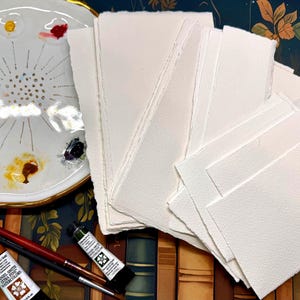May include: A selection of watercolour art supplies, including a white ceramic palette with paint, watercolour paper, paint tubes, and paint brushes. The paper is bright white and textured. The palette has a gold rim.