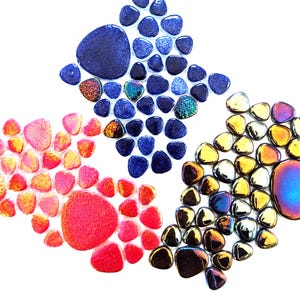 May include: A collection of small, smooth, rounded glass pebbles arranged in three distinct clusters. One group features deep blue and iridescent tones, another displays vibrant red and orange hues, and the third shows metallic gold and purple.