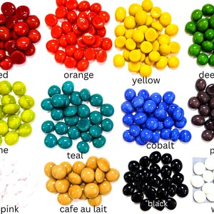 May include: A collection of small, smooth, rounded glass gems arranged in twelve distinct color groups labeled red, orange, yellow, deep green, lime, teal, cobalt, purple, pale pink, cafe au lait, black, and white on a white background.