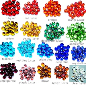 Puede incluir: Una colecci&oacute;n de peque&ntilde;as gemas de vidrio redondas con fondo plano dispuestas en veinte grupos de colores distintos, incluyendo rojo rub&iacute;, rojo lustre, naranja, naranja lustre, amarillo, amarillo lustre, verde profundo lustre, verde manzana, azul cerceta, azul cerceta lustre, azul brillante, azul profundo, azul profundo lustre, violeta, violeta lustre, marr&oacute;n, marr&oacute;n lustre y transparente lustre, cada uno etiquetado con su nombre.