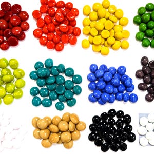 May include: A collection of smooth, round glass gems arranged in twelve distinct color groups including red, orange, yellow, green, lime, teal, blue, purple, white, tan, black, and a second white set, all featuring a flat base and a domed top.