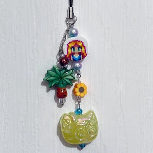 Puede incluir: Un amuleto amarillo iridiscente con un diseño de espiral, un amuleto de palmera verde, un amuleto de girasol amarillo y un amuleto blanco con una imagen pixelada de una niña con cabello rubio que lleva un vestido azul. Los amuletos están unidos a una cadena plateada con cuentas de perlas.