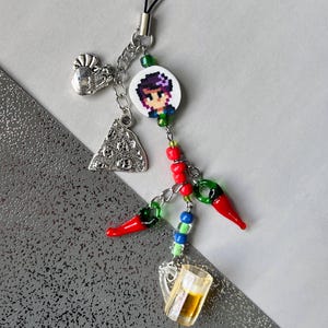 Puede incluir: Un abalorio colorido para teléfono con un personaje pixelado, una rebanada de pizza, chiles y una jarra de cerveza. El abalorio está unido a una cuerda negra.