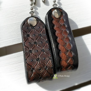 May include: Two brown leather fan pull chains with silver metal accents. One chain has a geometric pattern and the other has a braided pattern. The chains are hanging from a white surface. The text "3 Parks Design" is visible in the bottom right corner of the image.