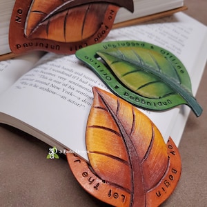 May include: Three leaf-shaped bookmarks made of leather in brown, green, and orange. Each bookmark features embossed veins and text along the border. The brown leaf reads No Stone Left Unturned, the green leaf reads Unplugged & Offline, and the orange leaf reads Let The Journey Begin.
