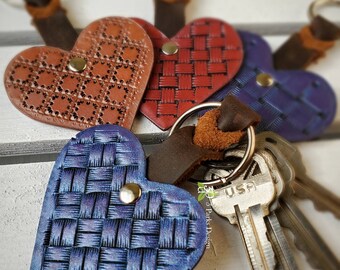May include: Three leather heart-shaped keychains with different designs. One is brown with a dotted pattern, one is red with a woven pattern, and one is blue with a woven pattern. The keychains are attached to a silver key ring with keys.