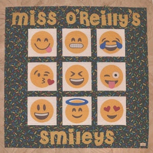 Pode incluir: Um cobertor preto e branco com um padrão de confete colorido. O cobertor tem uma faixa dourada na parte superior que diz "miss o'reilly's" e uma faixa dourada na parte inferior que diz "smileys". O cobertor é dividido em nove quadrados, cada um com um rosto sorridente amarelo diferente.