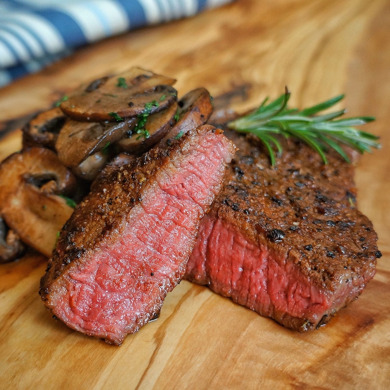 May include: A close-up of a cooked steak with a crispy crust and a juicy red interior. The steak is sliced in half and served with sauteed mushrooms and a sprig of rosemary.